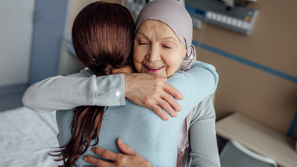 Jüngere Frau umarmt ältere Frau mit Kopftuch in einem Krankenhauszimmer. 
