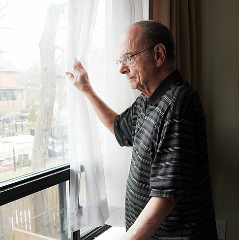 Ein älterer Mann zieht eine Gardine zur Seite und blickt aus dem Fenster